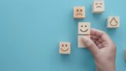 Hand choosing a happy face wooden block from a set of emojis on a blue background.